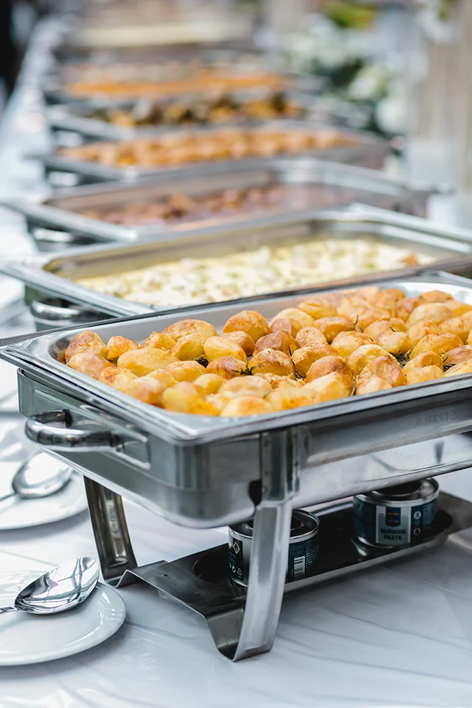 Ein Buffet mit verschiedenen Gerichten in Chafing Dishes aus Edelstahl. Die nächste Schale enthält Bratkartoffeln. Weitere Gerichte sind cremige und gegrillte Speisen. Löffel liegen neben den Tabletts auf einer weißen Tischdecke.