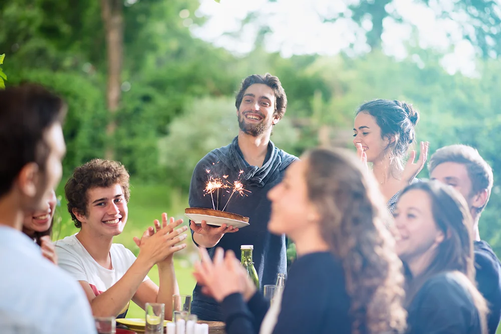 Eine Gruppe junger Erwachsener feiert im Freien. Eine lächelnde Person hält eine Torte mit Wunderkerzen in der Hand, während andere klatschen und jubeln. Die helle und üppige Atmosphäre lässt auf ein fröhliches, zwangloses Beisammensein in einem Garten oder Park schließen.