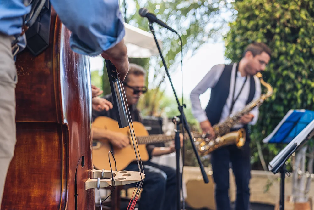 Eine Jazzband spielt im Freien. Ein Bassist spielt im Vordergrund einen Kontrabass, ein Gitarrist klimpert in der Mitte und ein Saxophonist spielt im Hintergrund. Bäume und Grünflächen sorgen für eine natürliche Kulisse.