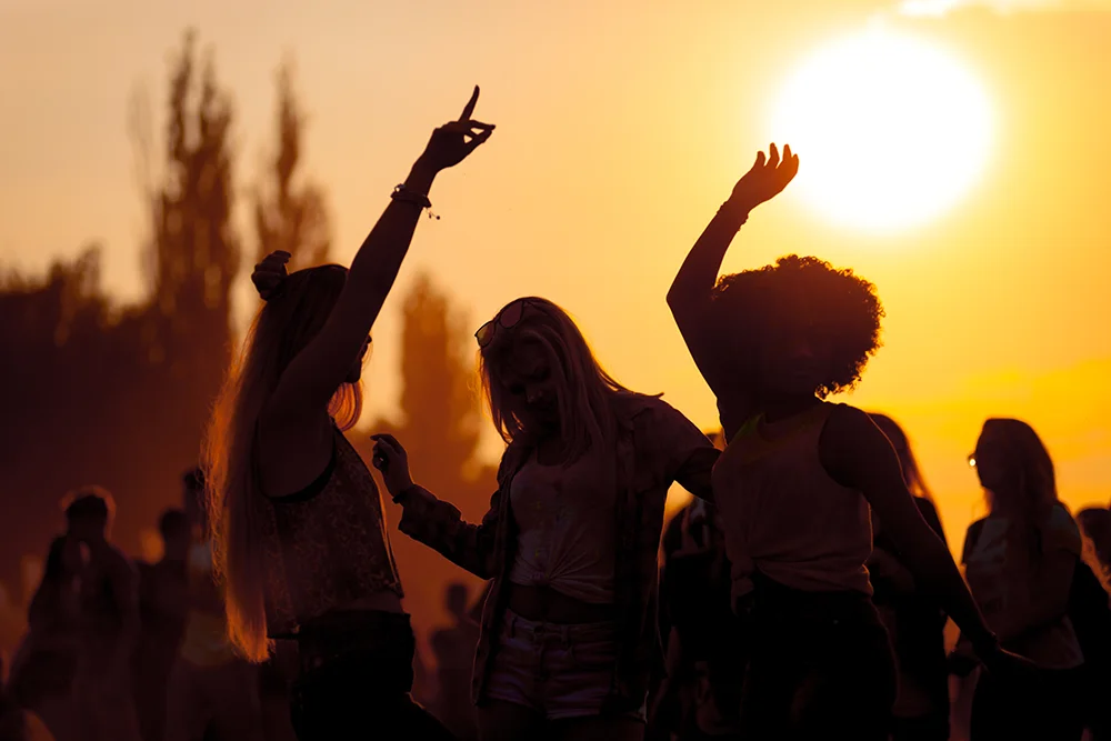 Silhouetten von drei Personen, die bei Sonnenuntergang energisch tanzen, mit einer Menschenmenge im Hintergrund. Der Himmel leuchtet in warmen Orangetönen und in der Ferne sind Bäume zu sehen.